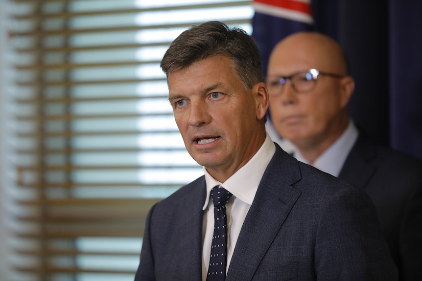 A man in a suit speaking, with another man out of focus behind him.