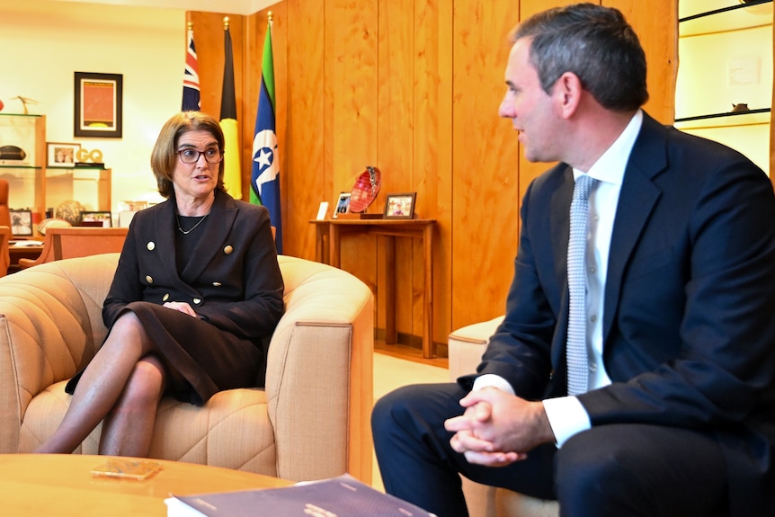 A woman with short brown hair and glasses sits in an armchair talking to a man in a suit.